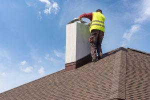 A roofer in a yellow vest stands on top of a brown shingle roof to perform an inspection.