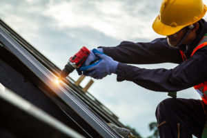 A roofer in a hardhat and vest uses a nail gun to install roofing shingles.